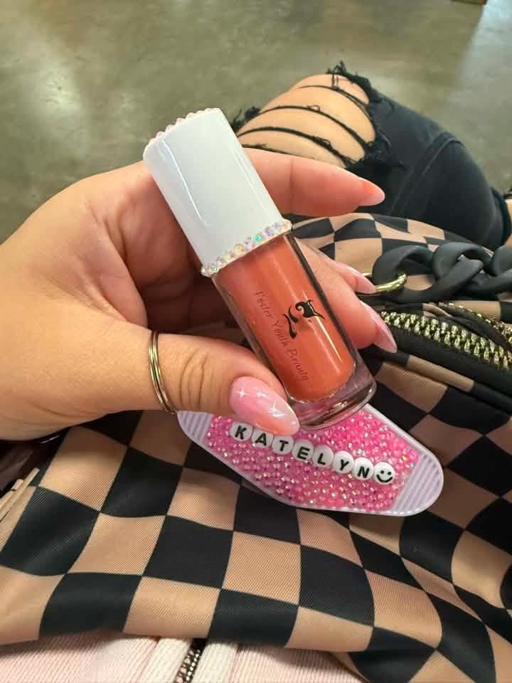Hand holding a coral-colored liquid lipstick with white cap and pink glittery name plate, displayed over black and beige checkered fabric