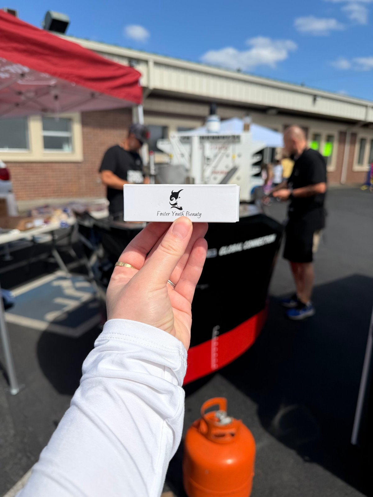Hand holding a business card at an outdoor event with food truck and people in background under clear blue sky