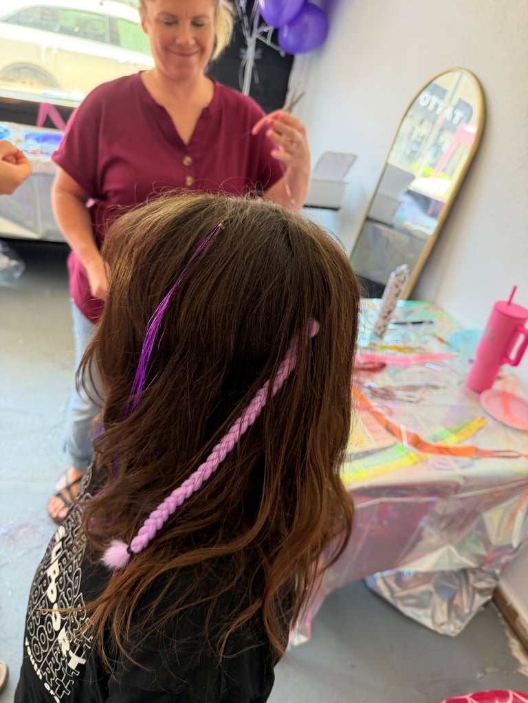Woman braiding pink and purple hair extensions into young girls long brown hair in a craft room setting