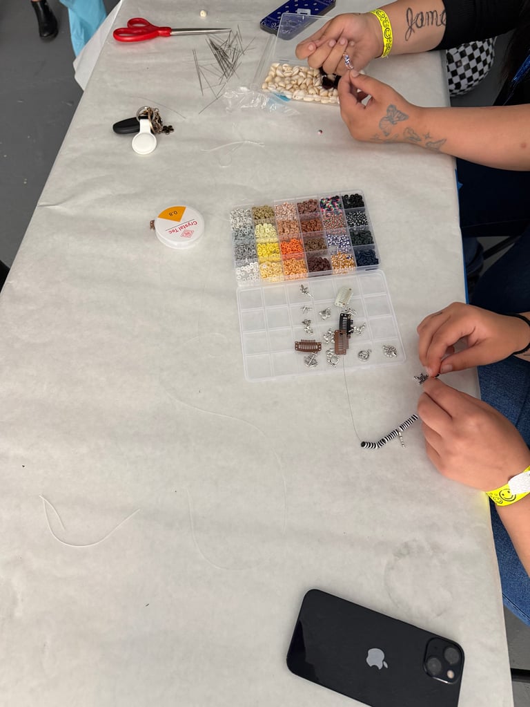 Three people working on beadwork craft project at table with colorful beads, tools, and smartphone