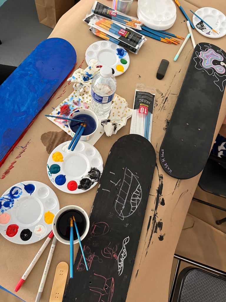 Art workspace on wooden table with skateboard decks, paint palettes, brushes, water cup, and art supplies